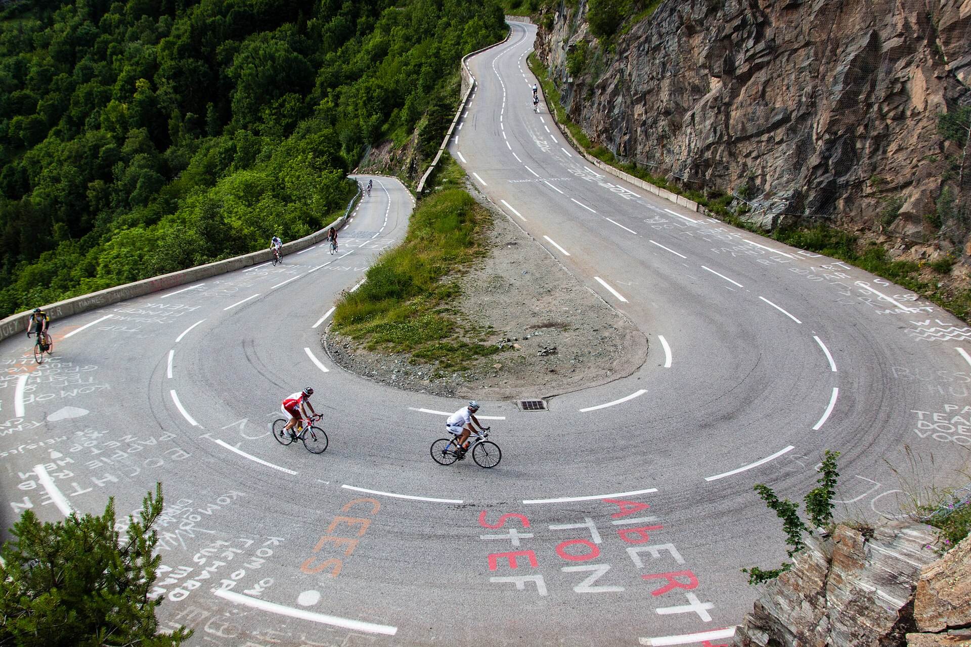 Deux cyclistes prennent un virage en épingle sur une route couverte d'inscriptions peintes, falaise et forêt autour