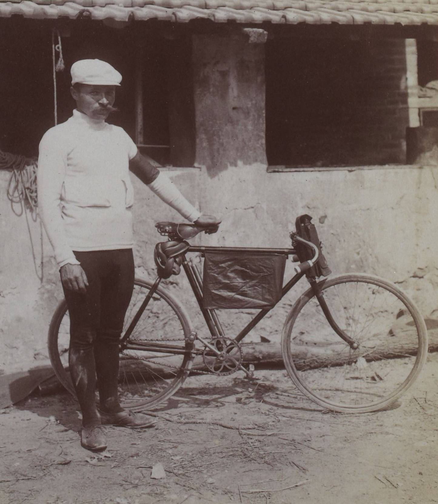 Maurice Garin, en maillot clair et casquette, pose debout à côté de sa bicyclette devant un bâtiment