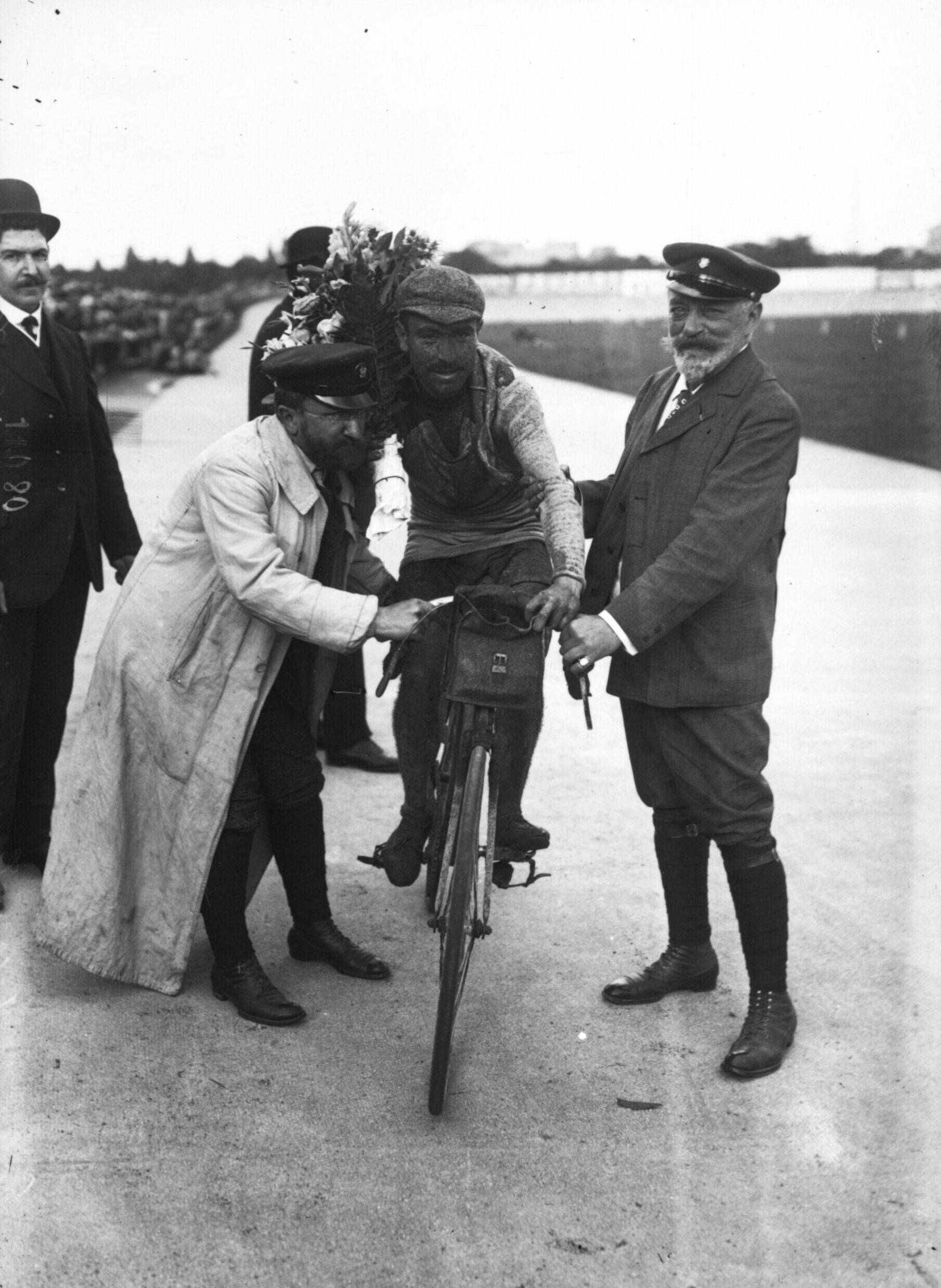 Octave Lapize sur sa bicyclette, entouré de trois hommes, un bouquet de fleurs derrière lui