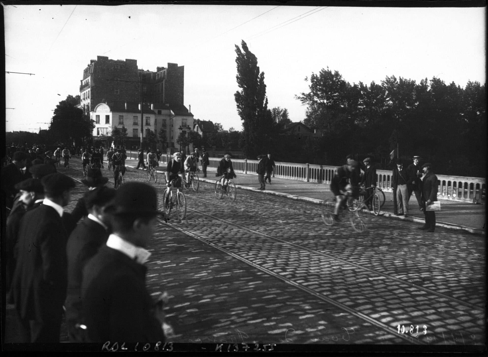 Coureurs sur une route pavée qui franchit un pont, des spectateurs en chapeau sur le bord
