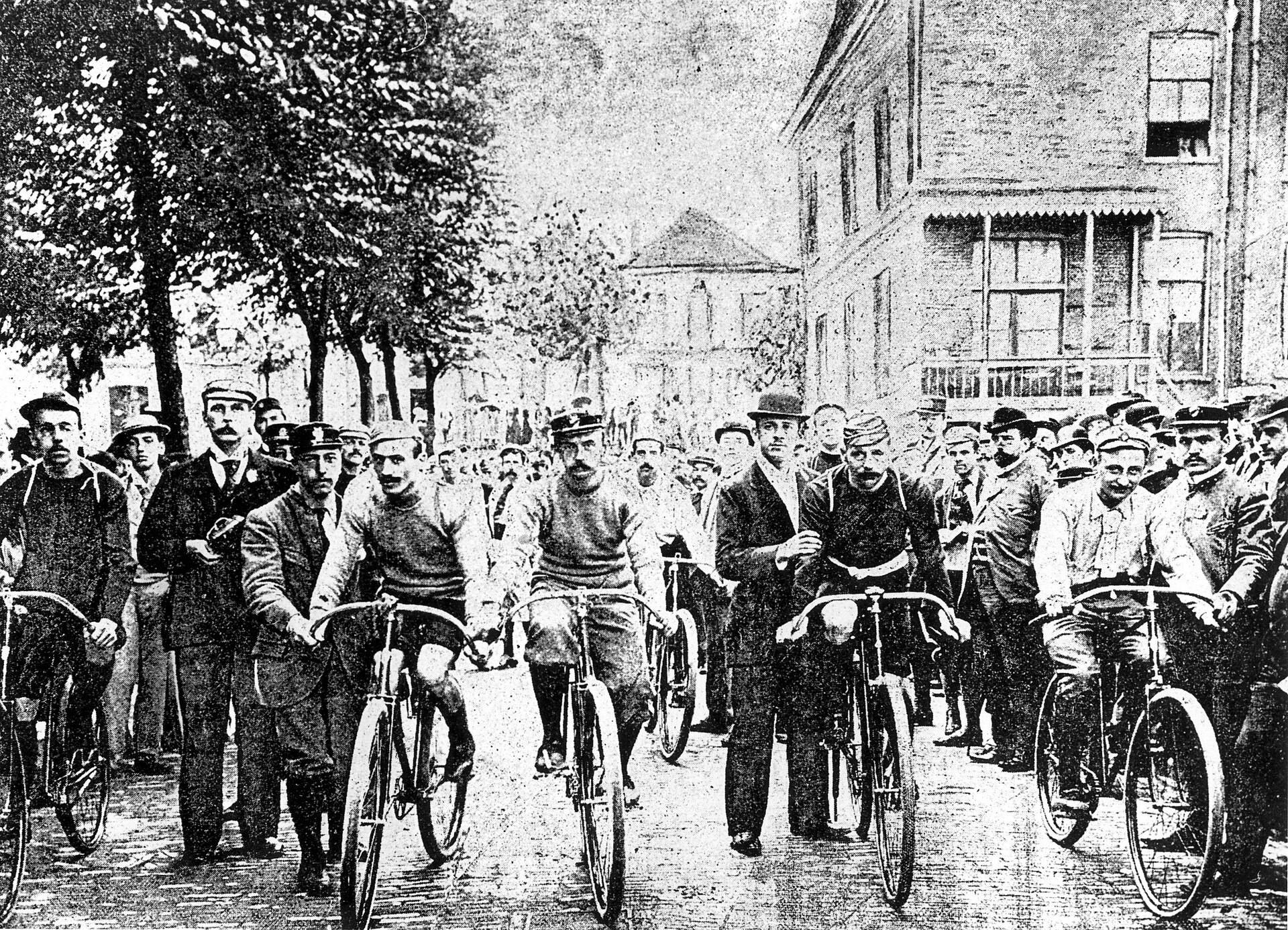 Groupe de coureurs tenant leurs bicyclettes, entourés de spectateurs, dans une rue bordée d'arbres