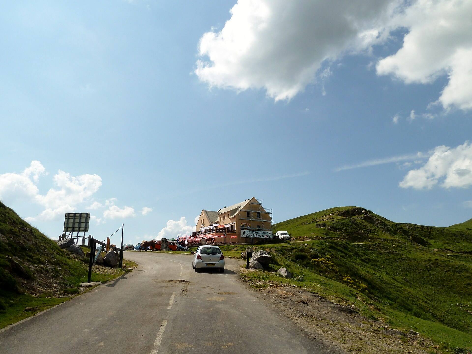 Route de montagne étroite qui monte entre des pâturages vers un bâtiment au sommet du col d'Aubisque, voiture blanche sur la chaussée