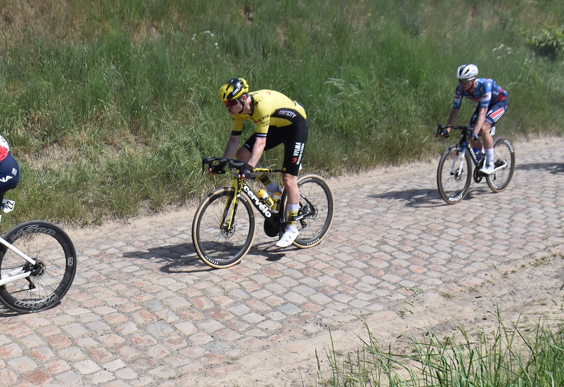 Trois coureurs sur un secteur pavé bordé d'herbe haute