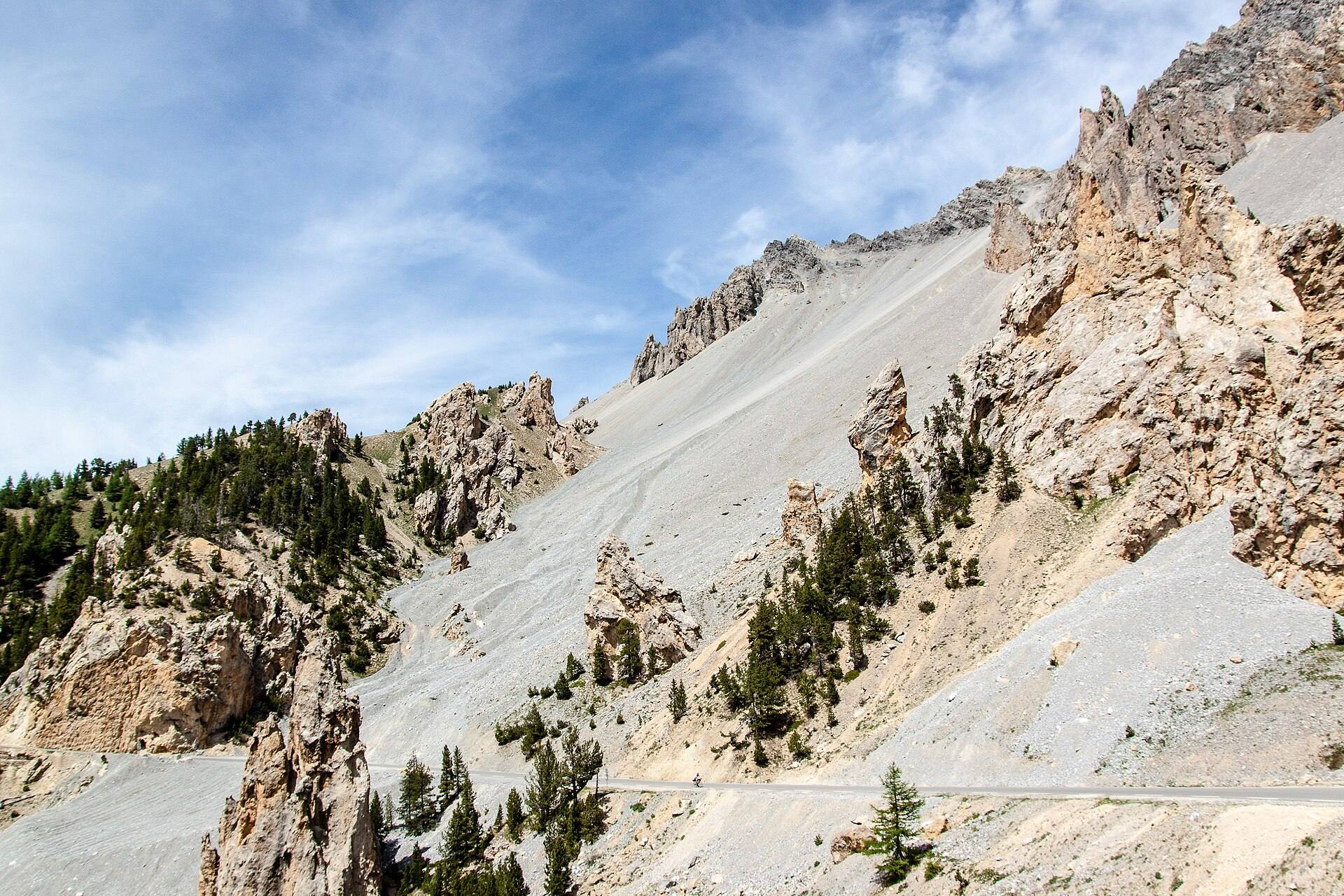 Pentes d'éboulis gris et aiguilles de roche ocre, quelques mélèzes, la route qui traverse le pierrier en bas de l'image