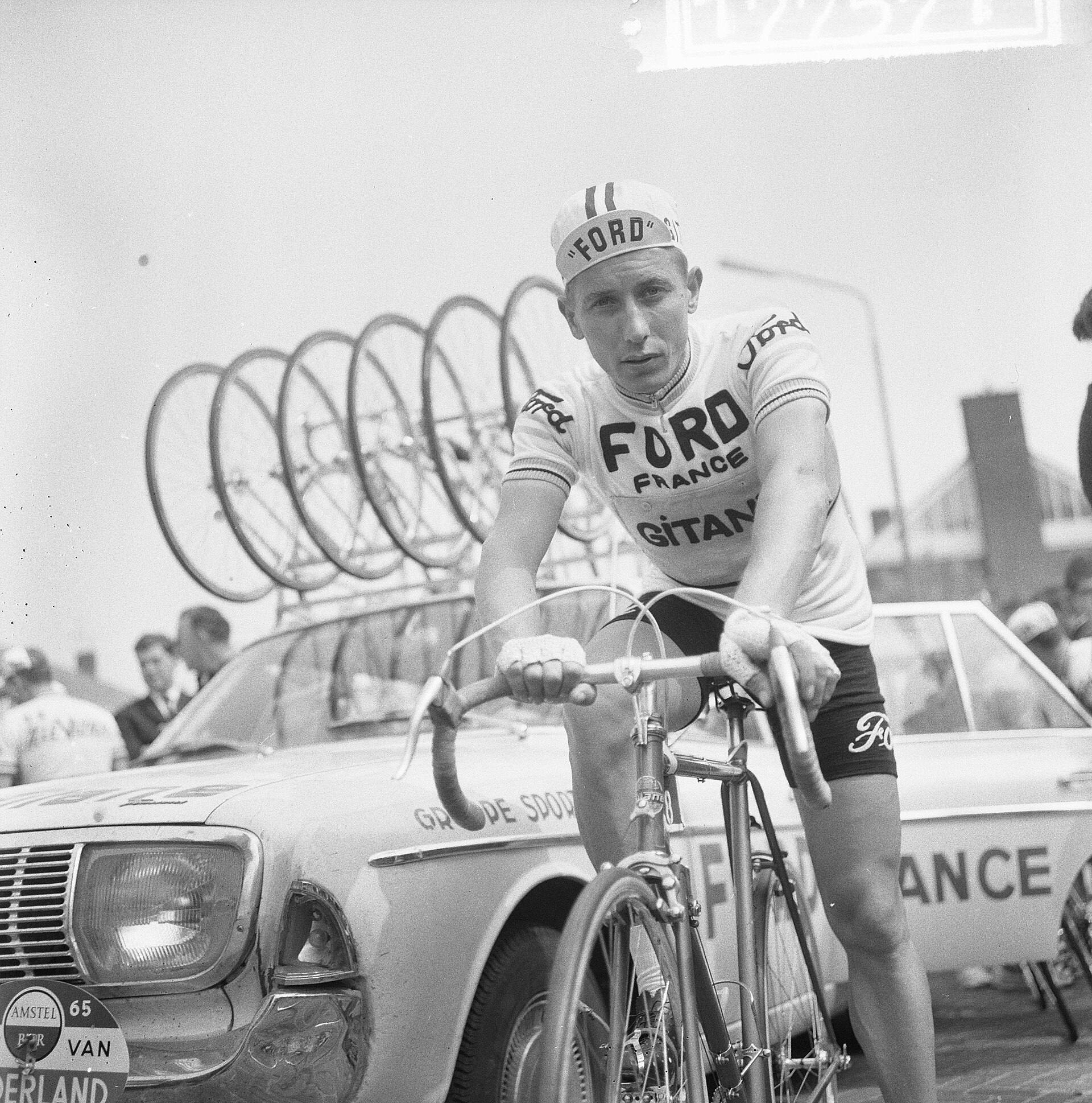 Un coureur blond en maillot « Ford France Gitane » et casquette « Ford », assis sur sa bicyclette, devant une voiture d'équipe « Ford France » chargée de roues sur le toit.