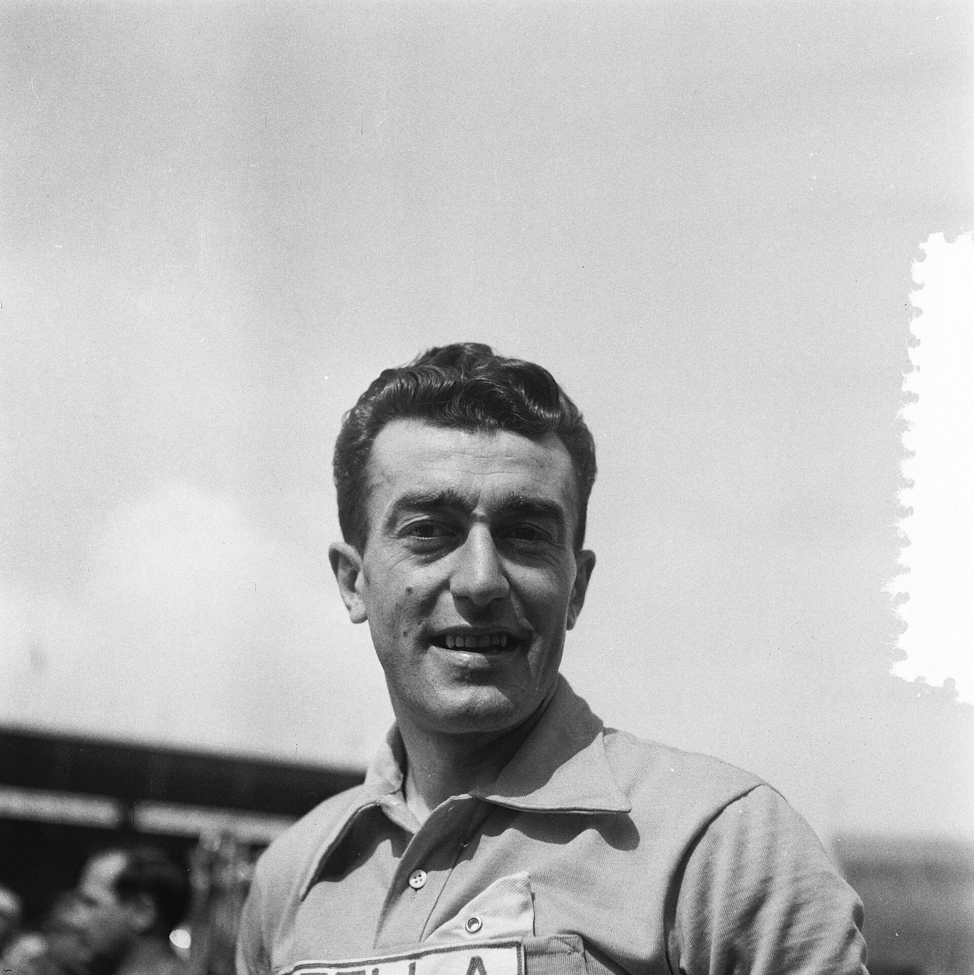 Gros plan d'un coureur souriant aux cheveux bruns ondulés, en maillot à col où l'on devine l'inscription « Stella », devant les tribunes d'un stade.