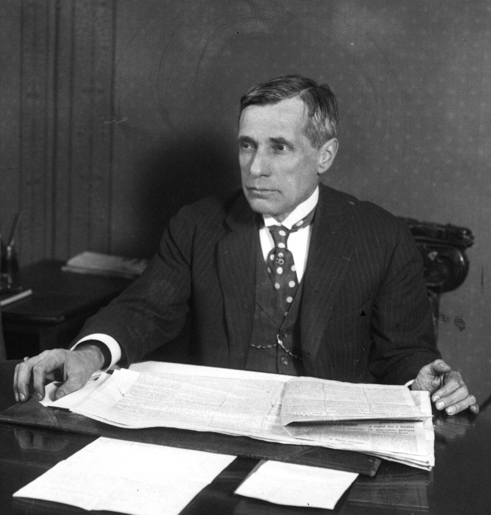 Un homme aux cheveux gris, en costume rayé, gilet et cravate à pois, assis à un bureau couvert de journaux dépliés, regarde vers la droite.