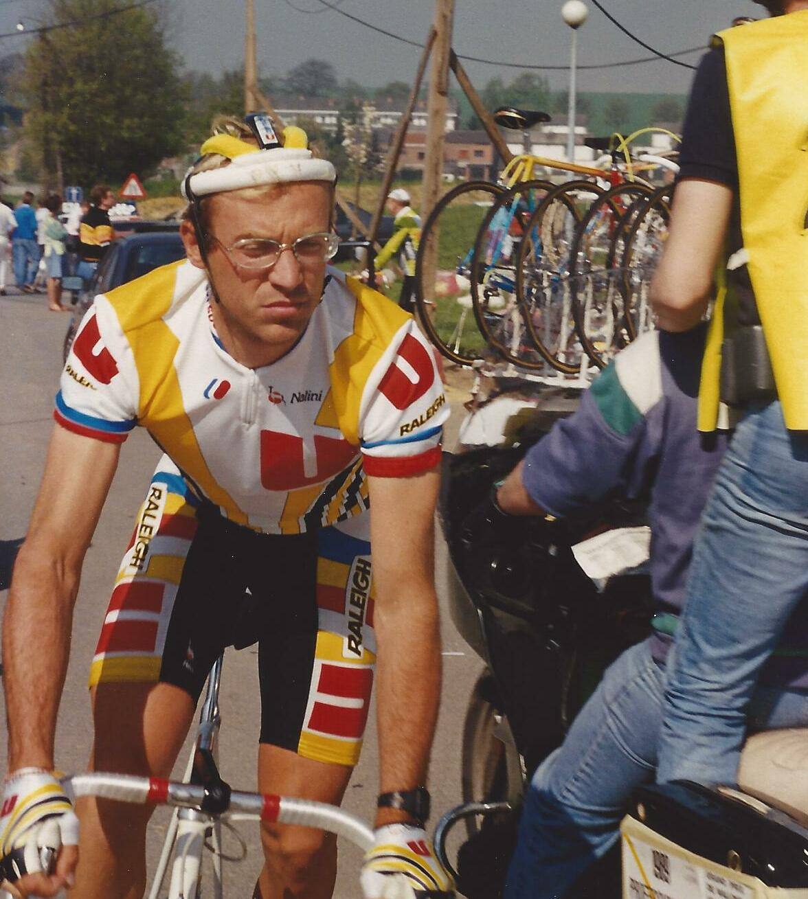 Photo en couleur d'un coureur blond à lunettes et bandeau, en maillot blanc, jaune et rouge orné de grands « U », sur sa bicyclette à côté d'un râtelier de vélos et d'une personne en gilet jaune.