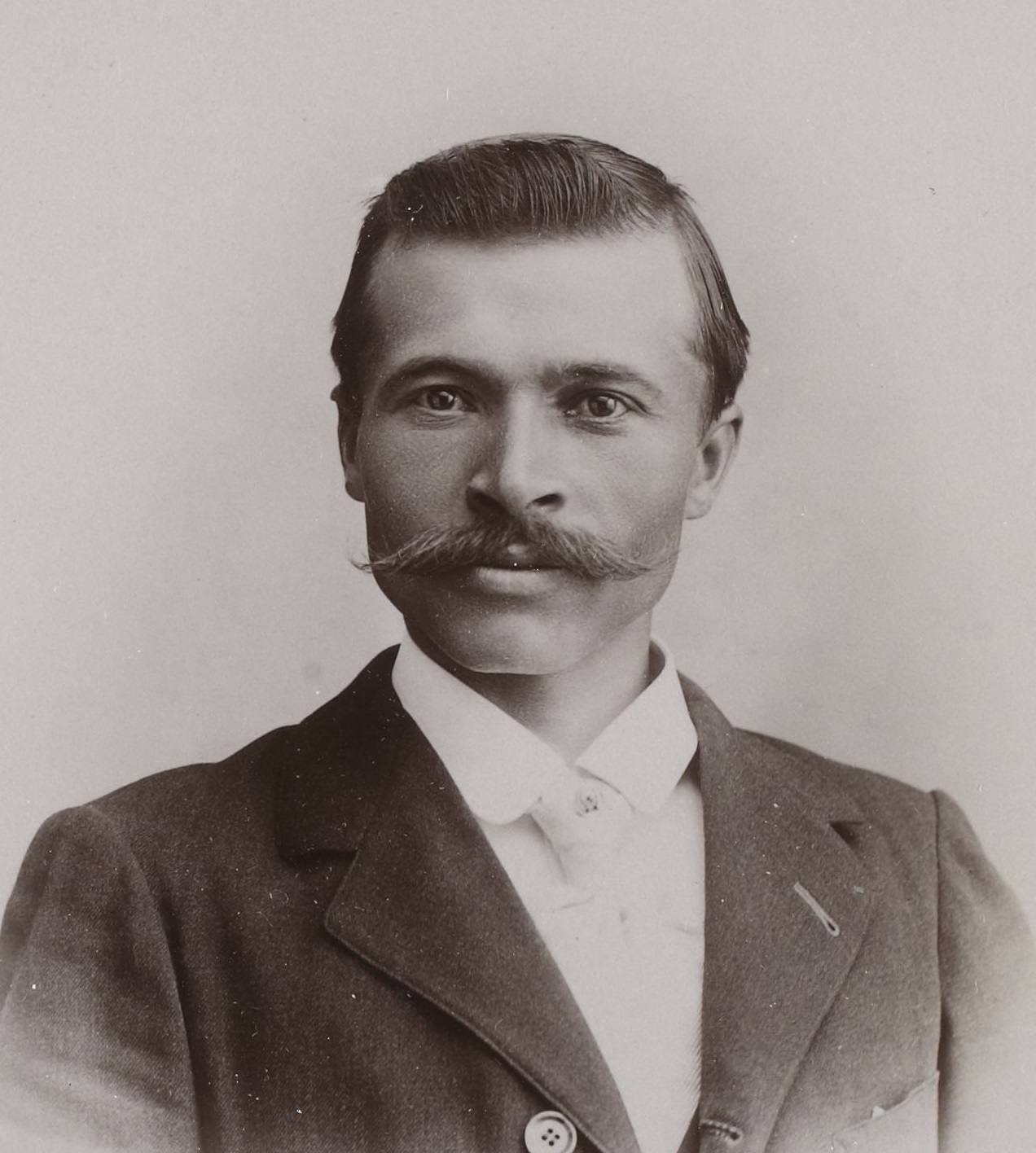 Portrait en buste, en tons sépia, d'un homme à grande moustache, cheveux courts coiffés en arrière, en veste sombre et chemise blanche, sur fond clair.