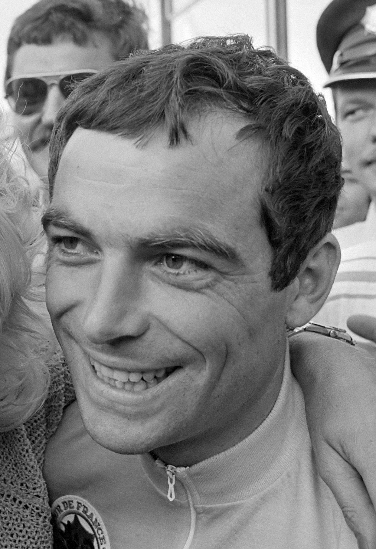 Gros plan d'un homme brun aux cheveux courts, souriant largement, en maillot zippé portant un écusson rond « Tour de France » en partie visible ; des visages coupés l'entourent.