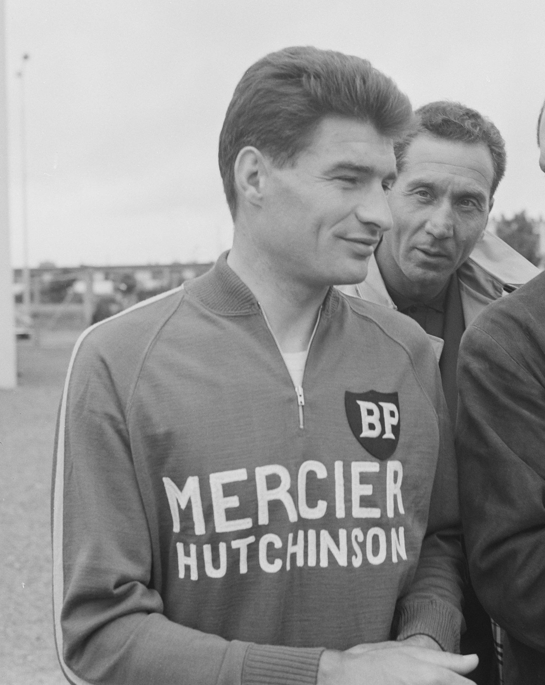 Portrait d'un coureur aux cheveux bruns, légèrement souriant, en veste zippée « BP Mercier Hutchinson », avec un homme derrière lui.