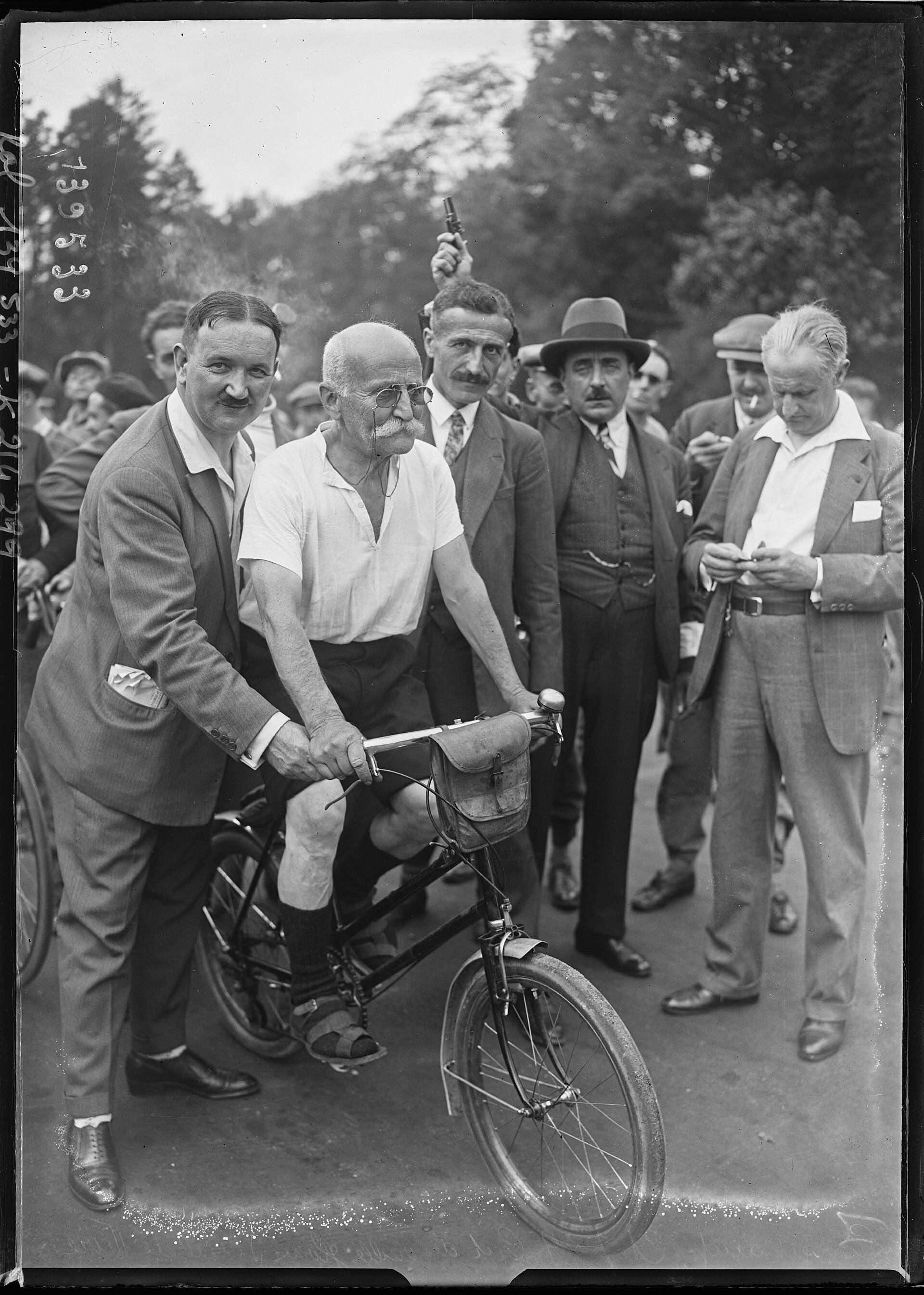 Un vieil homme chauve à moustache blanche et lunettes rondes, en chemise blanche, culotte sombre et sandales, est assis sur une bicyclette à sacoche de guidon ; un homme en costume le retient, d'autres l'entourent et l'un d'eux lève un pistolet.